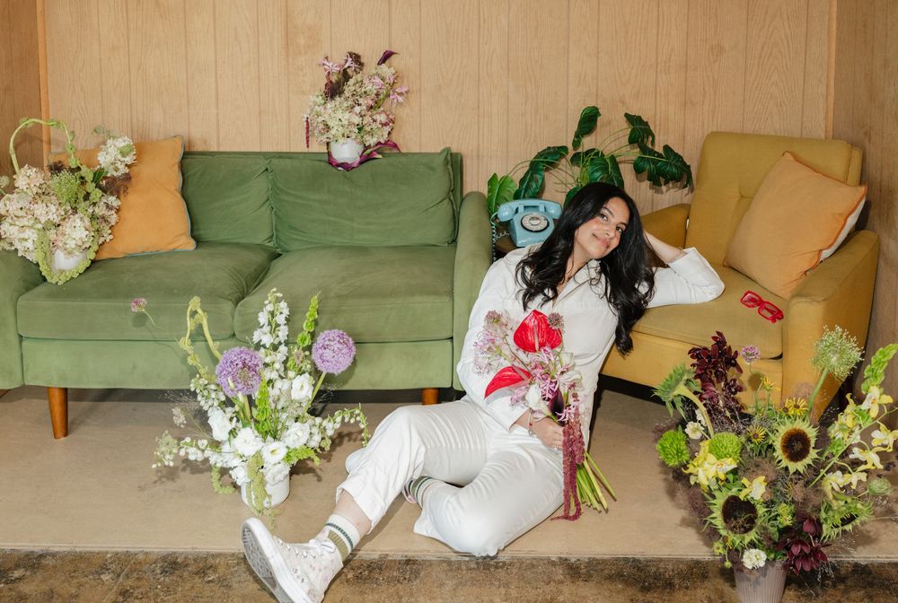 Woman sitting on floor surrounded by colorful floral arrangements near green sofas in a cozy room.
