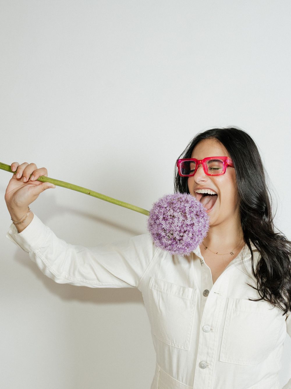 Woman in white jacket and pink glasses playfully pretending to lick a purple allium flower on a stem.