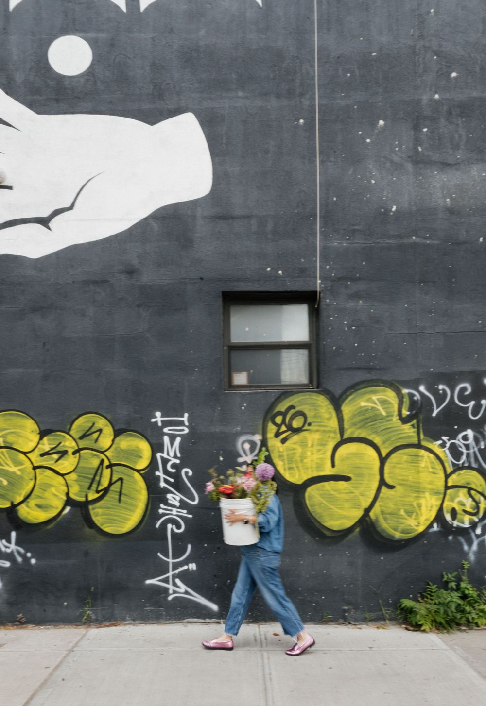 Person in denim carrying flowers walks past a gray wall with yellow graffiti and a large mural in an urban setting.