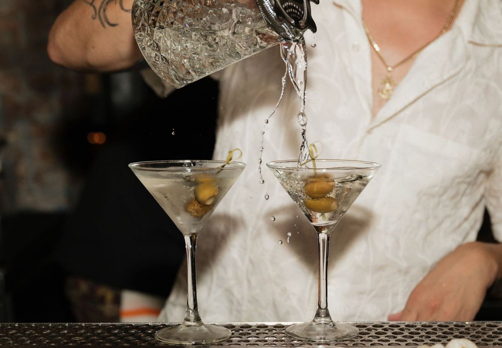 Bartender pouring dirty martini cocktails into two elegant glasses garnished with green olives.