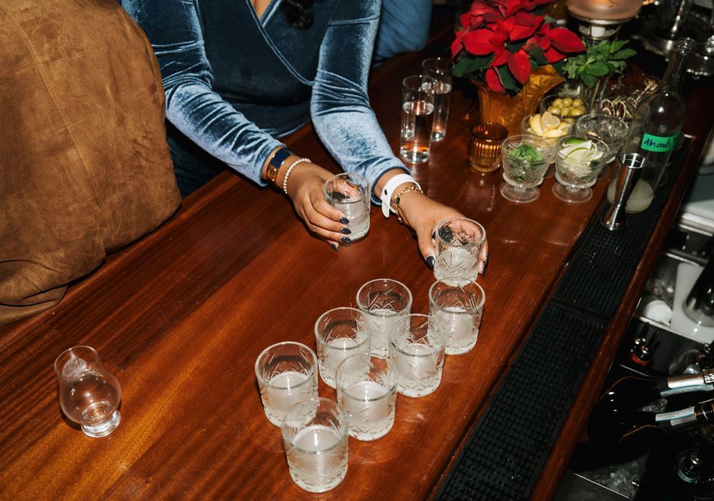 Bartender arranging multiple glass tumblers on a wooden bar counter in a dimly lit cocktail lounge.