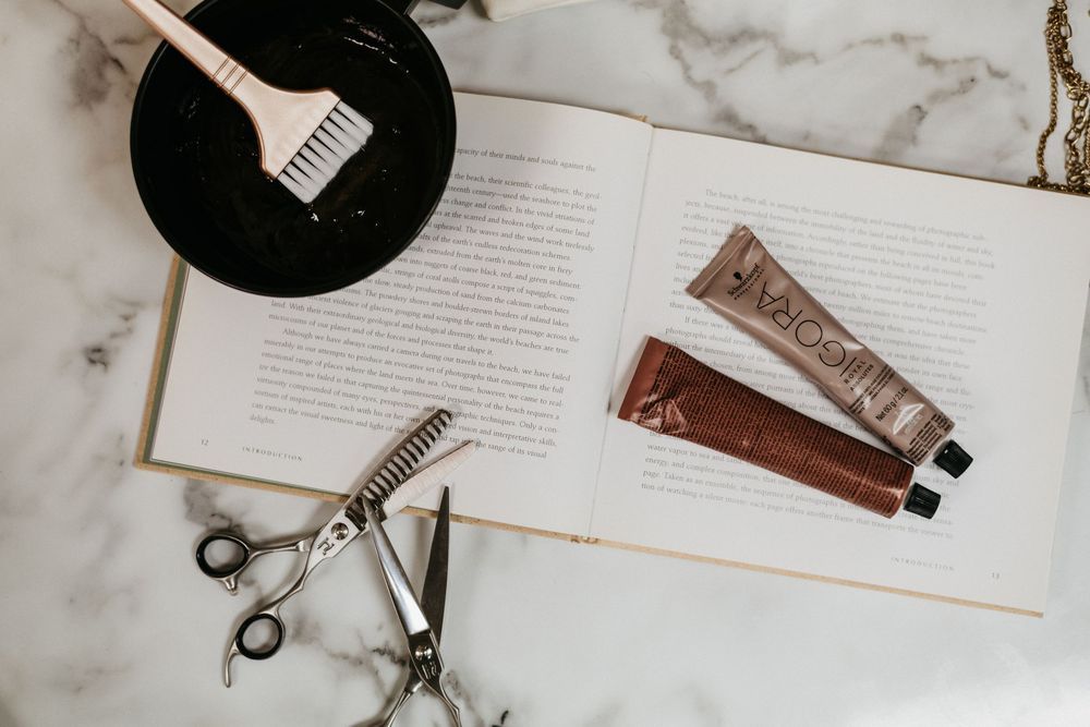 Hair coloring tools including scissors, dye tubes, and a tinting brush on an open book atop a marble surface.