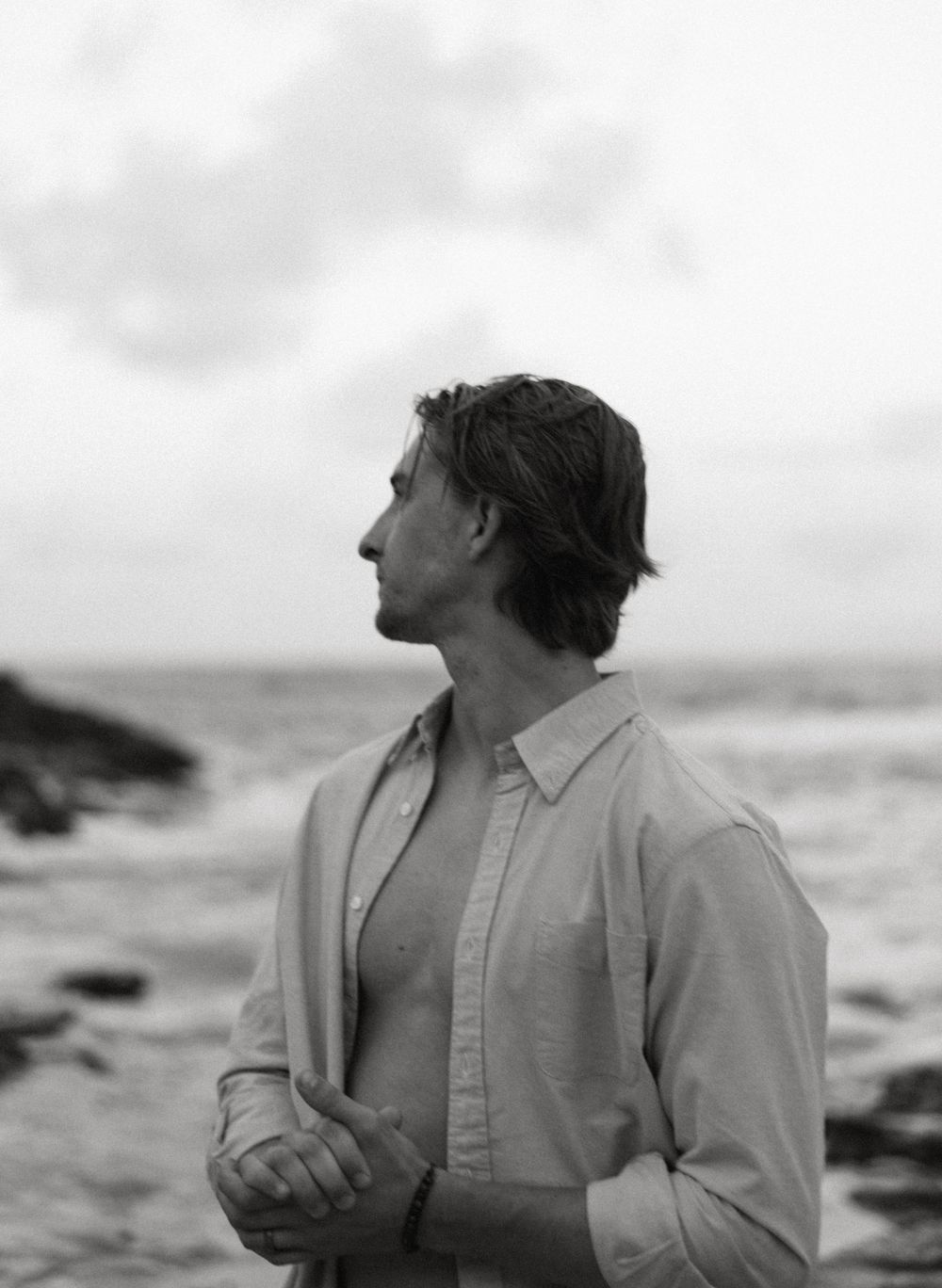 Black and white photo of a contemplative man with medium-length hair standing by a rocky ocean shore.