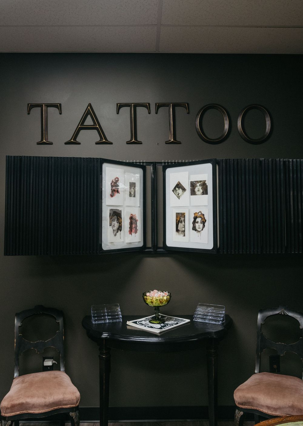 Dark tattoo parlor waiting area with portfolio books, two vintage chairs, and bold TATTOO lettering on wall.