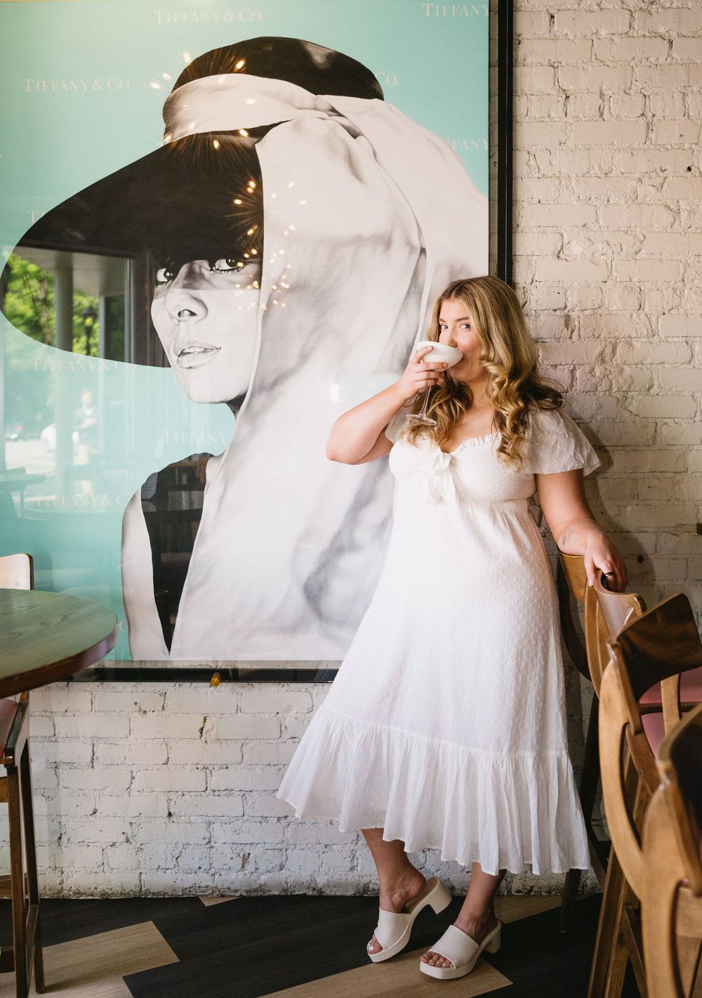 Woman in white midi dress sipping coffee in front of Audrey Hepburn poster at a chic cafe.