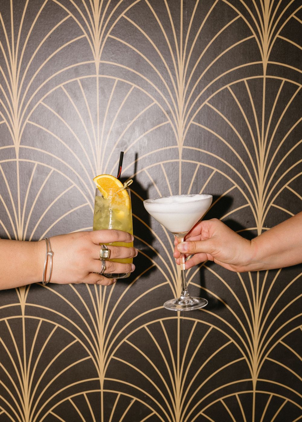 Two hands toasting with cocktails against an Art Deco gold and black wallpaper backdrop.