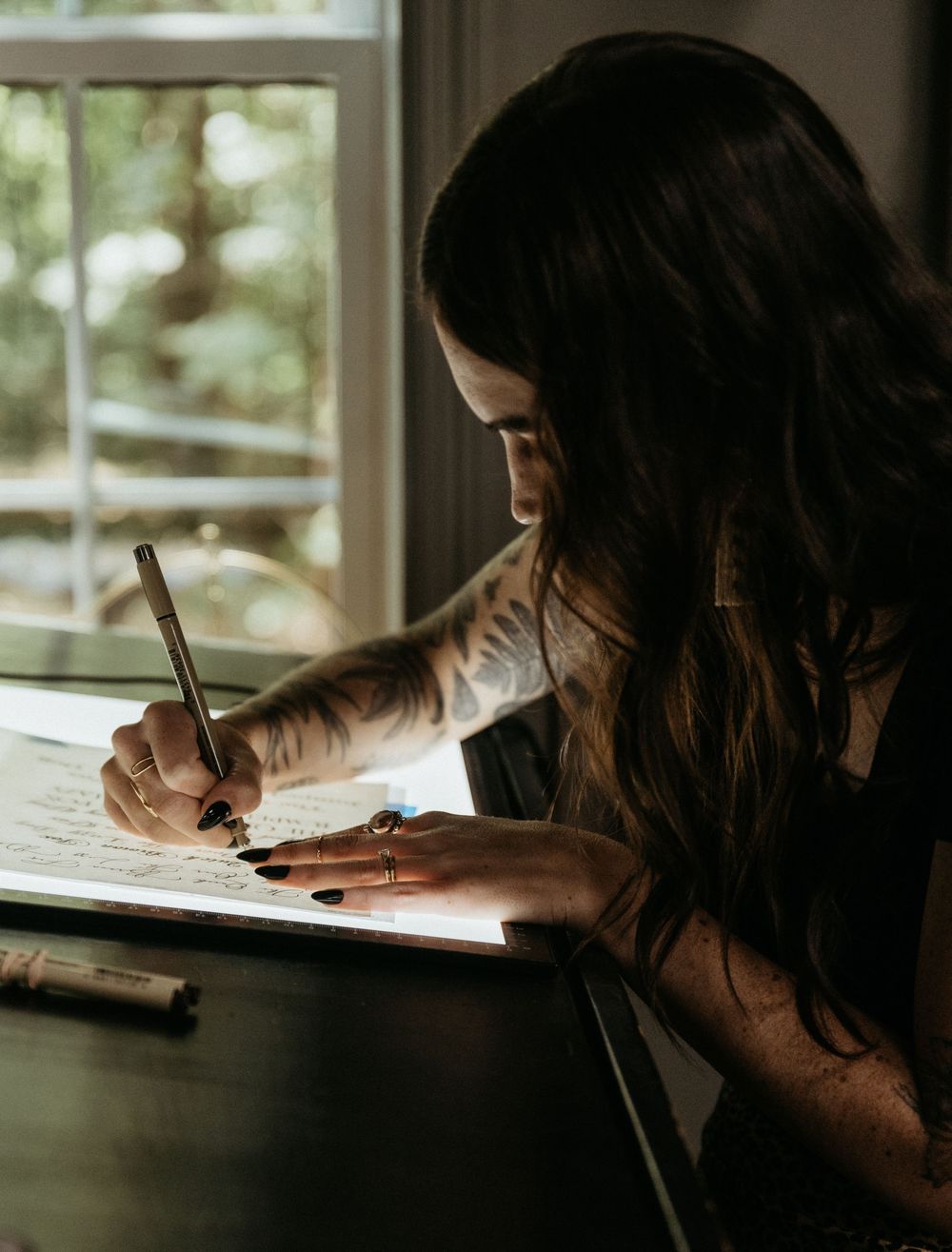 Woman with tattoos writing calligraphy at a desk near a window, focused on her artwork with a pen.