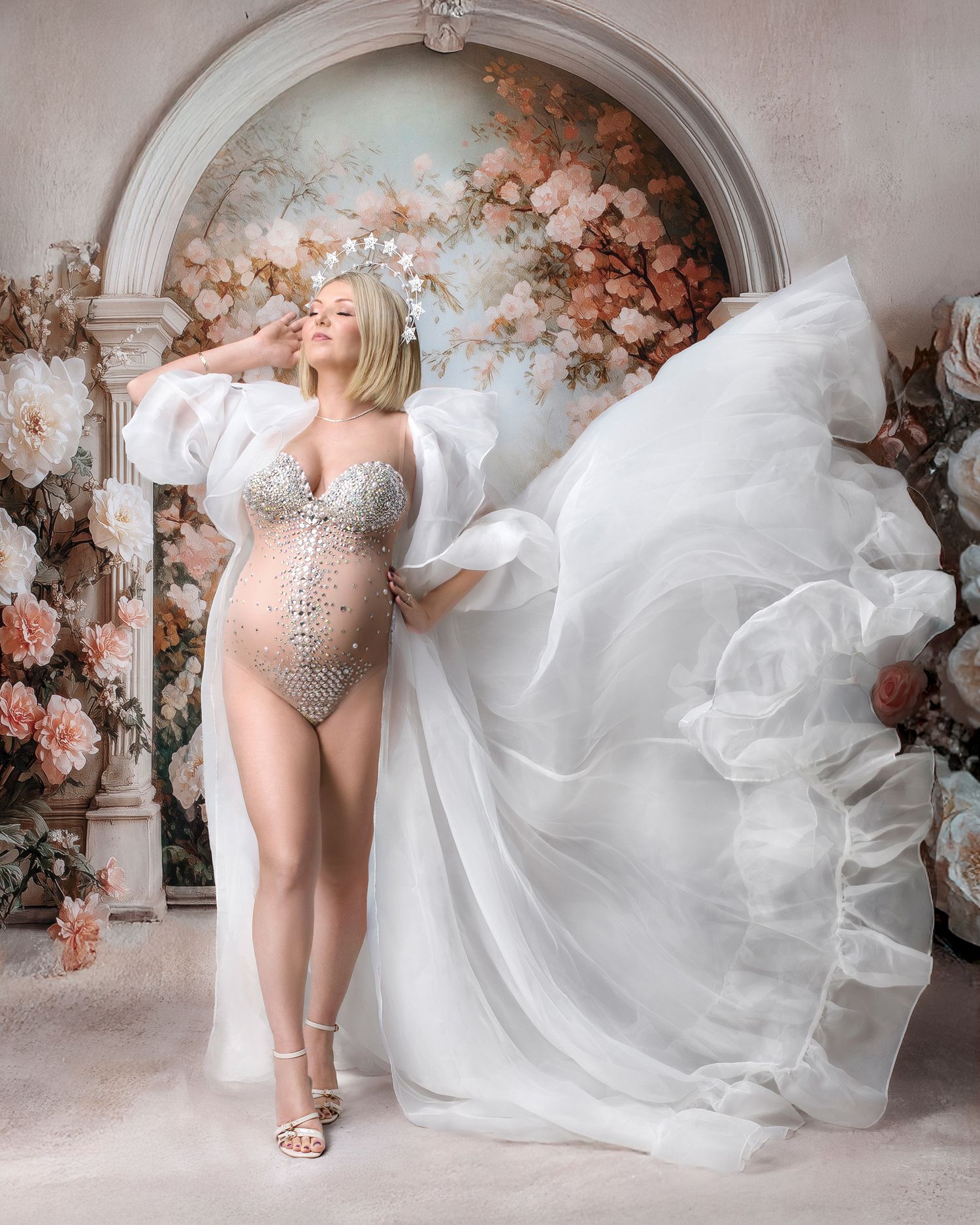 Ethereal pregnancy photo woman against gorgeous pastel coloured floral arch with her white robe flying up in the breeze.