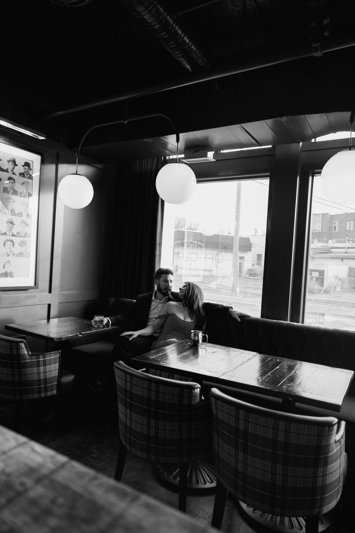 Black and white interior of a restaurant with plaid upholstered chairs, round pendant lights and large windows.