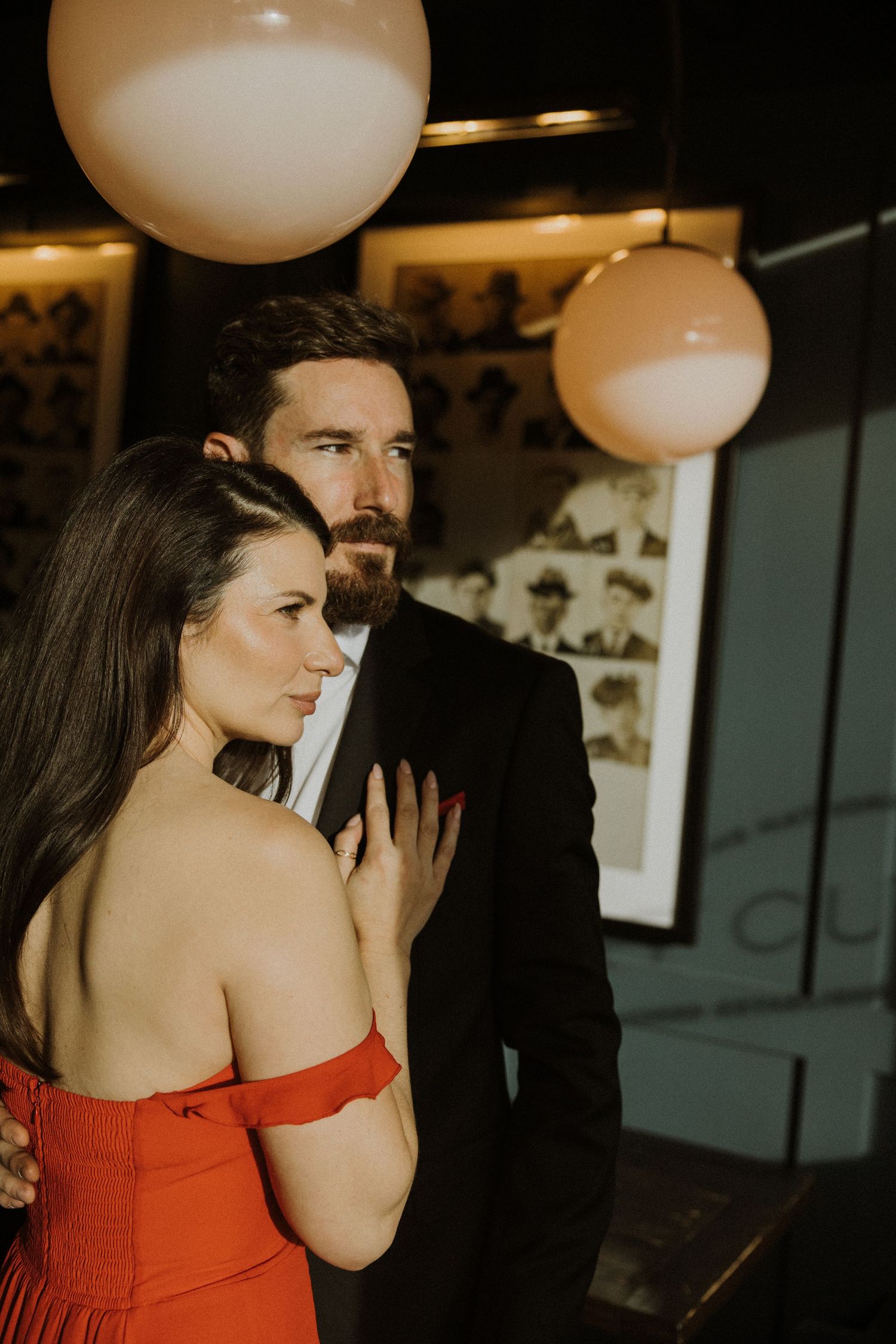 A couple shares an intimate moment in formal attire under round pendant lights in a dark venue.