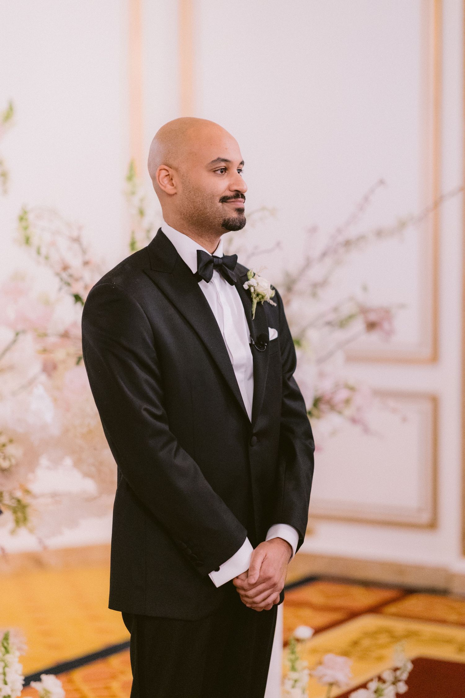 A groom in a classic black tuxedo and bow tie smiles emotionally during a wedding ceremony with cherry blossoms in background.