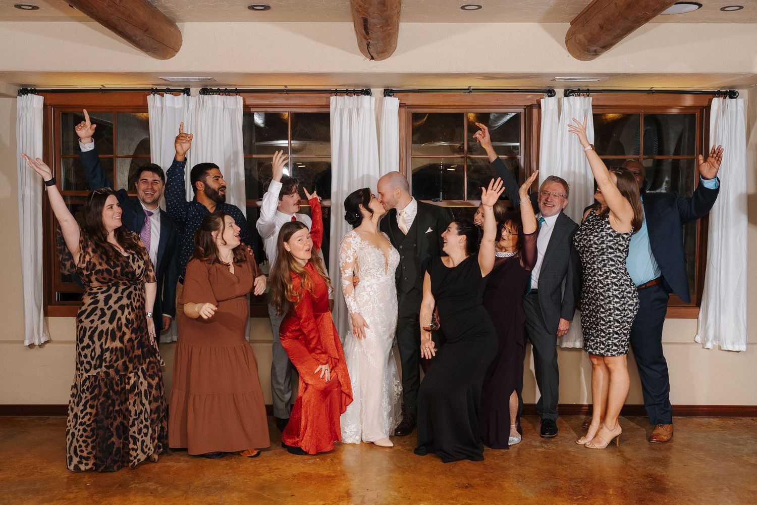 A group of wedding guests dancing and celebrating together in a rustic indoor venue with exposed wooden beams.
