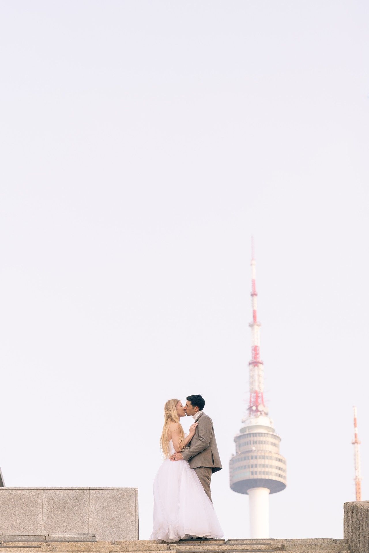 A romantic couple embraces in front of a white domed building with spires against a light-colored sky.
