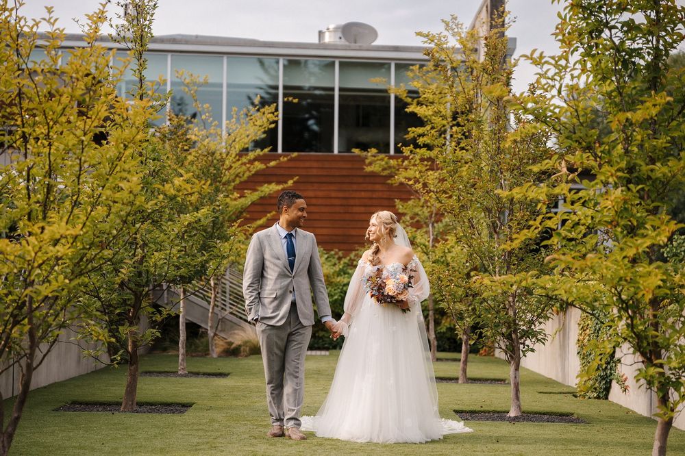 A couple in wedding attire walks together through a scenic garden setting with autumn trees.