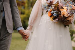 A couple holds hands while walking together outdoors, with the bride carrying a fall-themed floral bouquet.