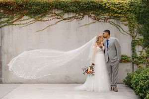 Elegant wedding couple poses against a stone wall with flowing veil and ivy creating a romantic garden atmosphere.