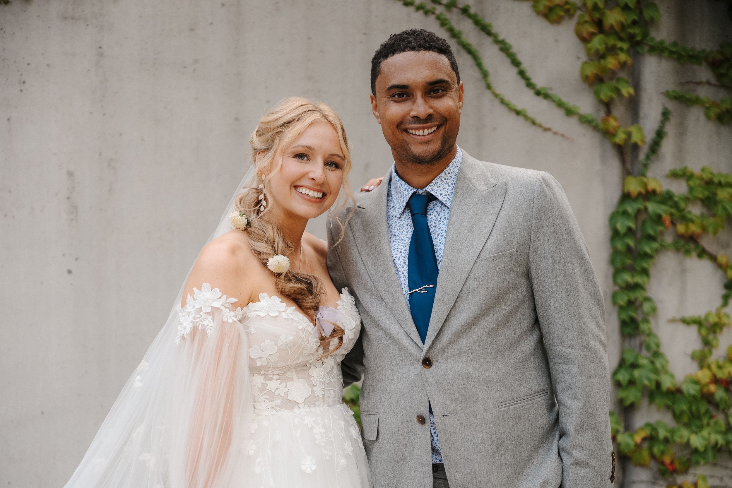 A wedding couple poses in front of an ivy-covered wall in formal attire during their outdoor photo session.