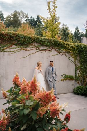 Couple poses against concrete wall with climbing vines and pink flowering plants in outdoor garden setting.