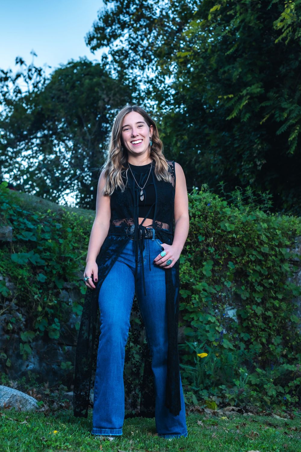 Person wearing a black sleeveless top and flared blue jeans poses outdoors against green foliage.