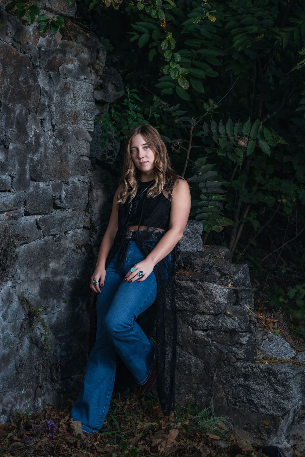 Moody portrait against stone wall with greenery features casual outfit of black top and flared blue jeans.