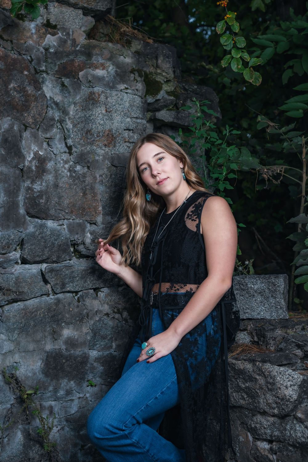 A black lace sleeveless top and blue jeans outfit photographed against a stone wall with green foliage in the background.