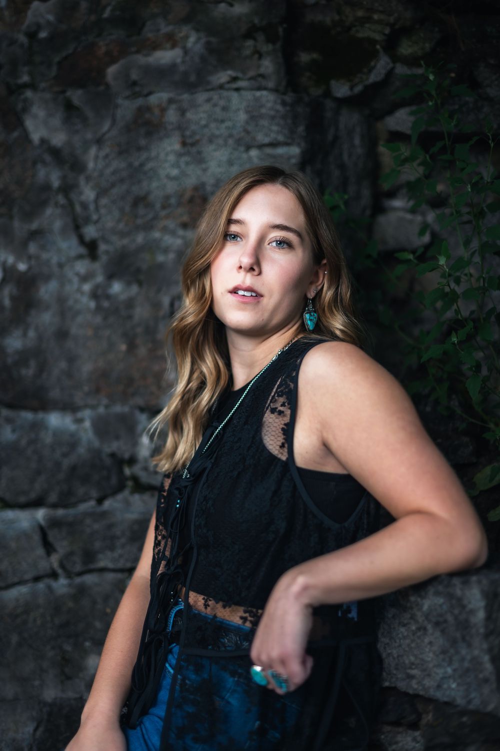 A woman in a black sleeveless top and denim overalls poses against a dark stone wall with dramatic lighting.