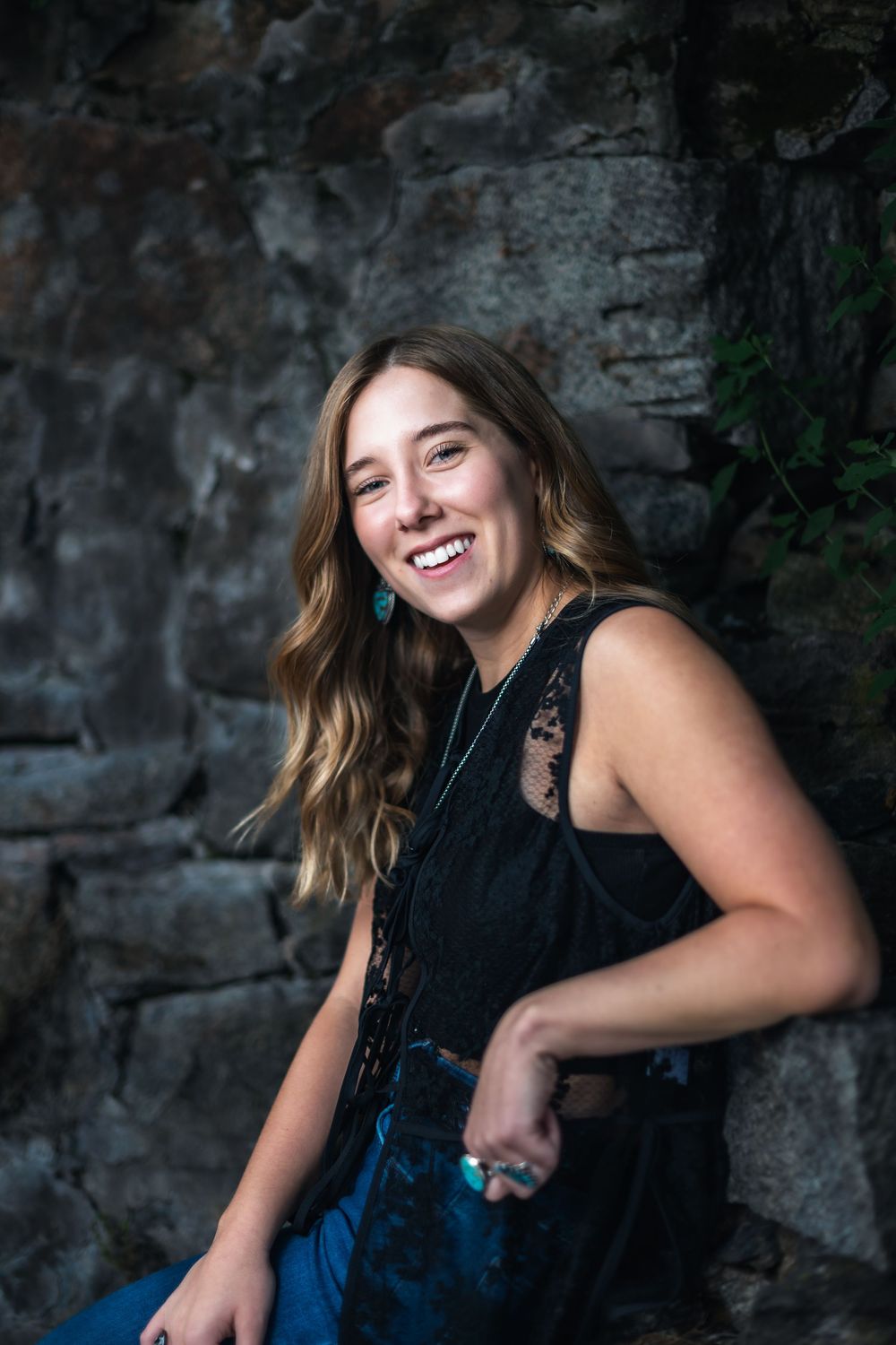 A person in a black top and denim smiles joyfully against a dark stone wall background.