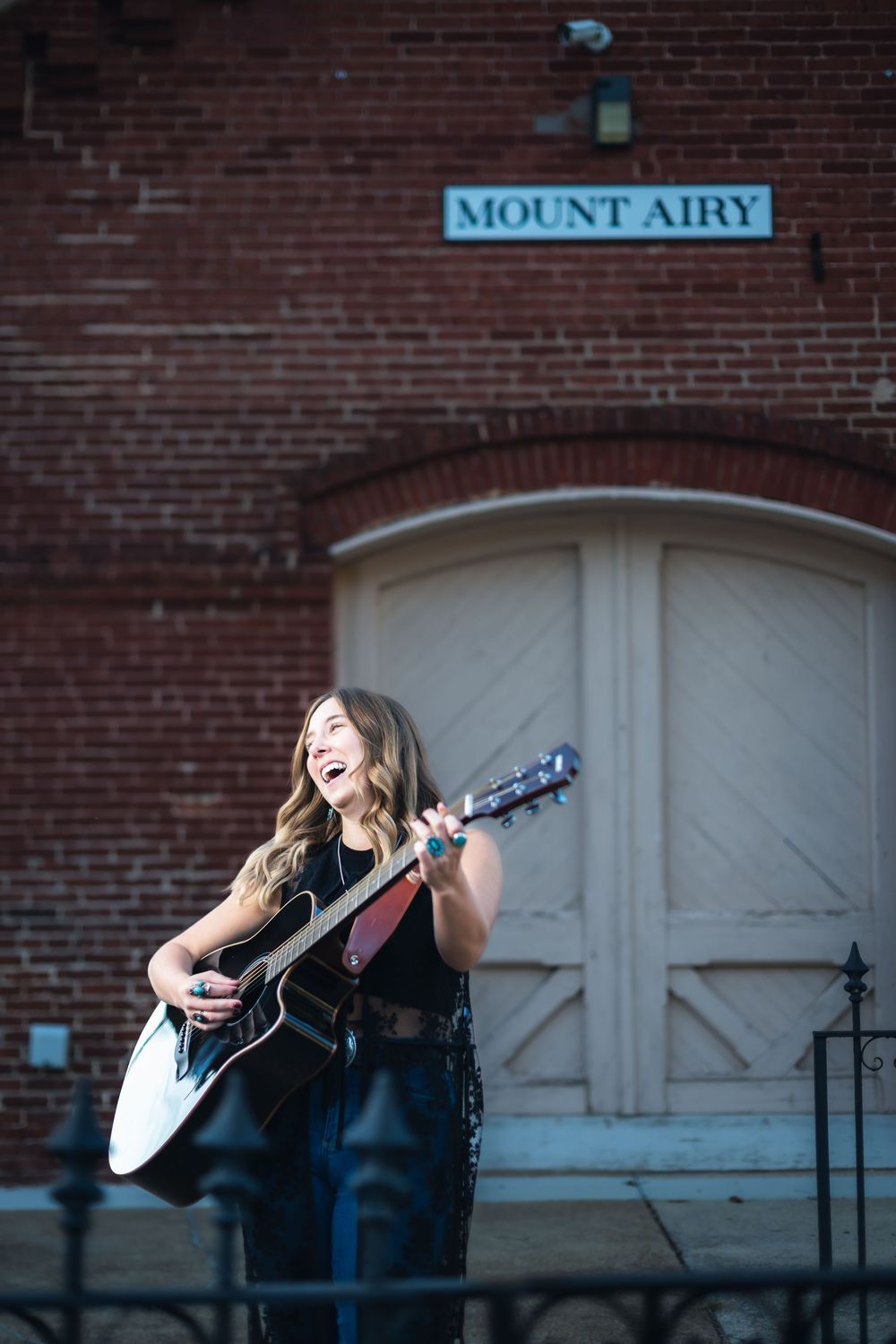 Musician performs with acoustic guitar outside brick building with Mount Airy sign in vintage urban setting. Hannah Ponce