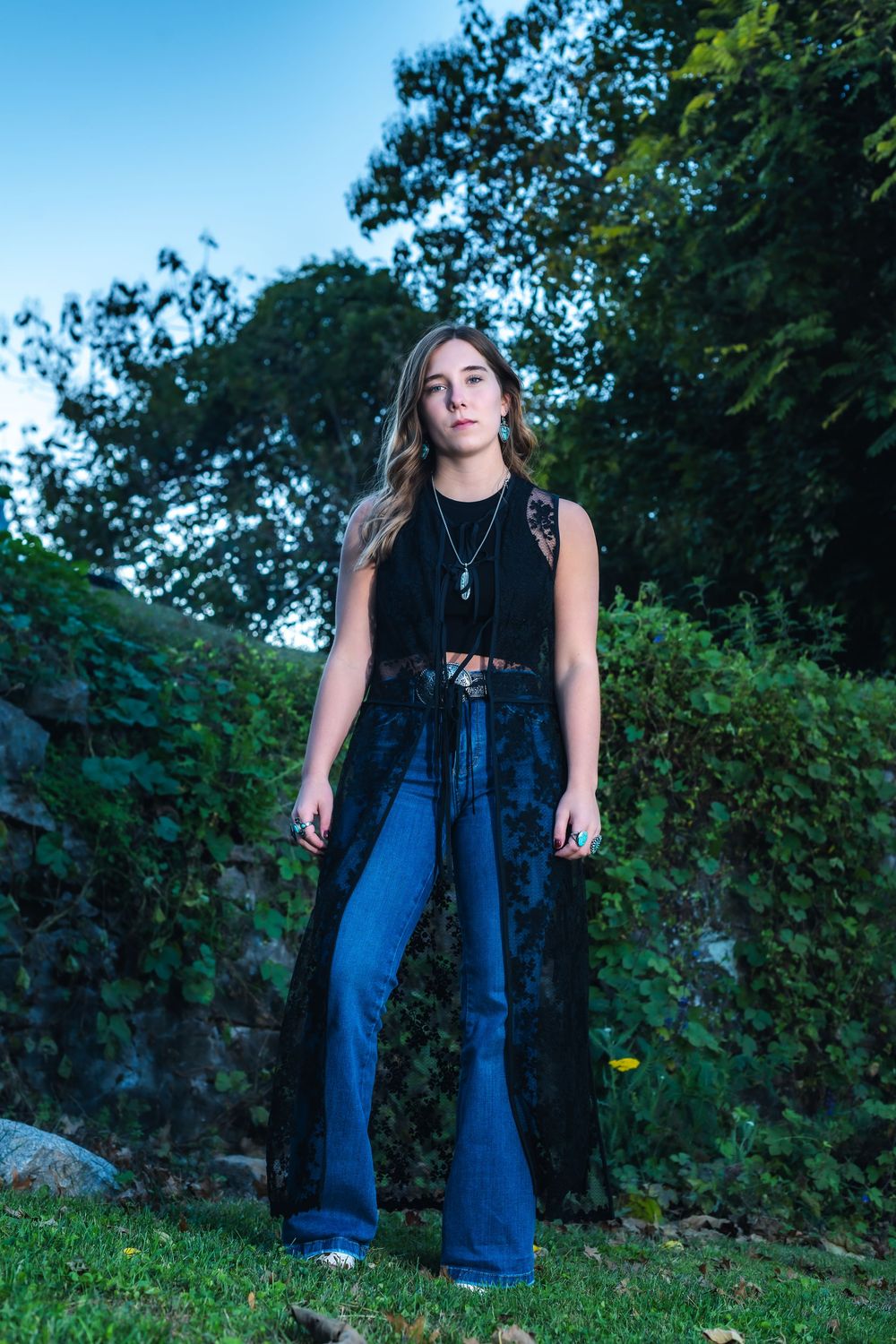 A person in a black sleeveless top and flared jeans poses in an outdoor garden setting with lush greenery.