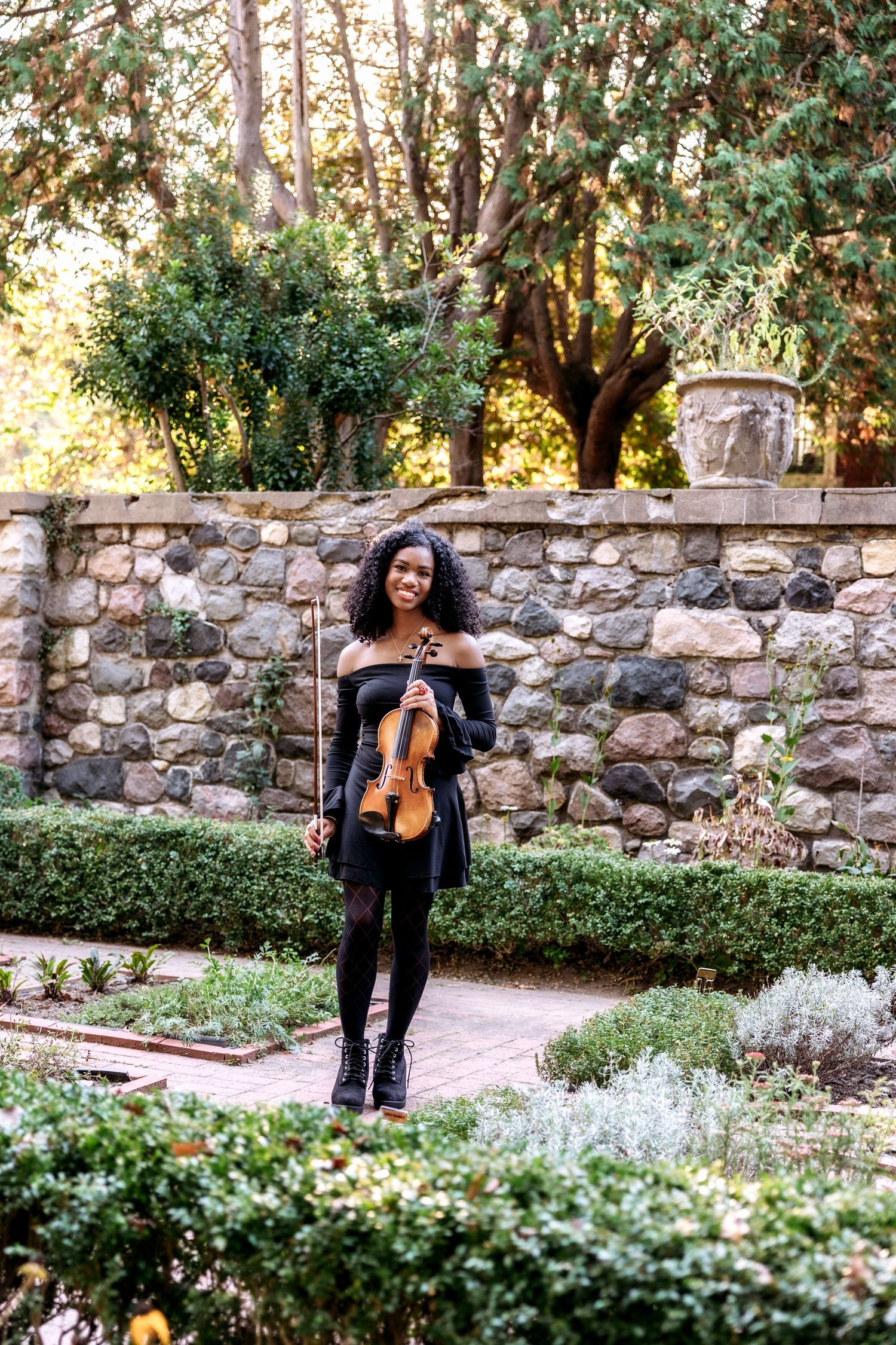 A violinist performs in a serene garden setting with stone walls and lush greenery in the background.