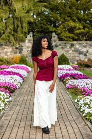 A garden pathway lined with vibrant pink and white flowers features a burgundy top and white maxi skirt outfit.