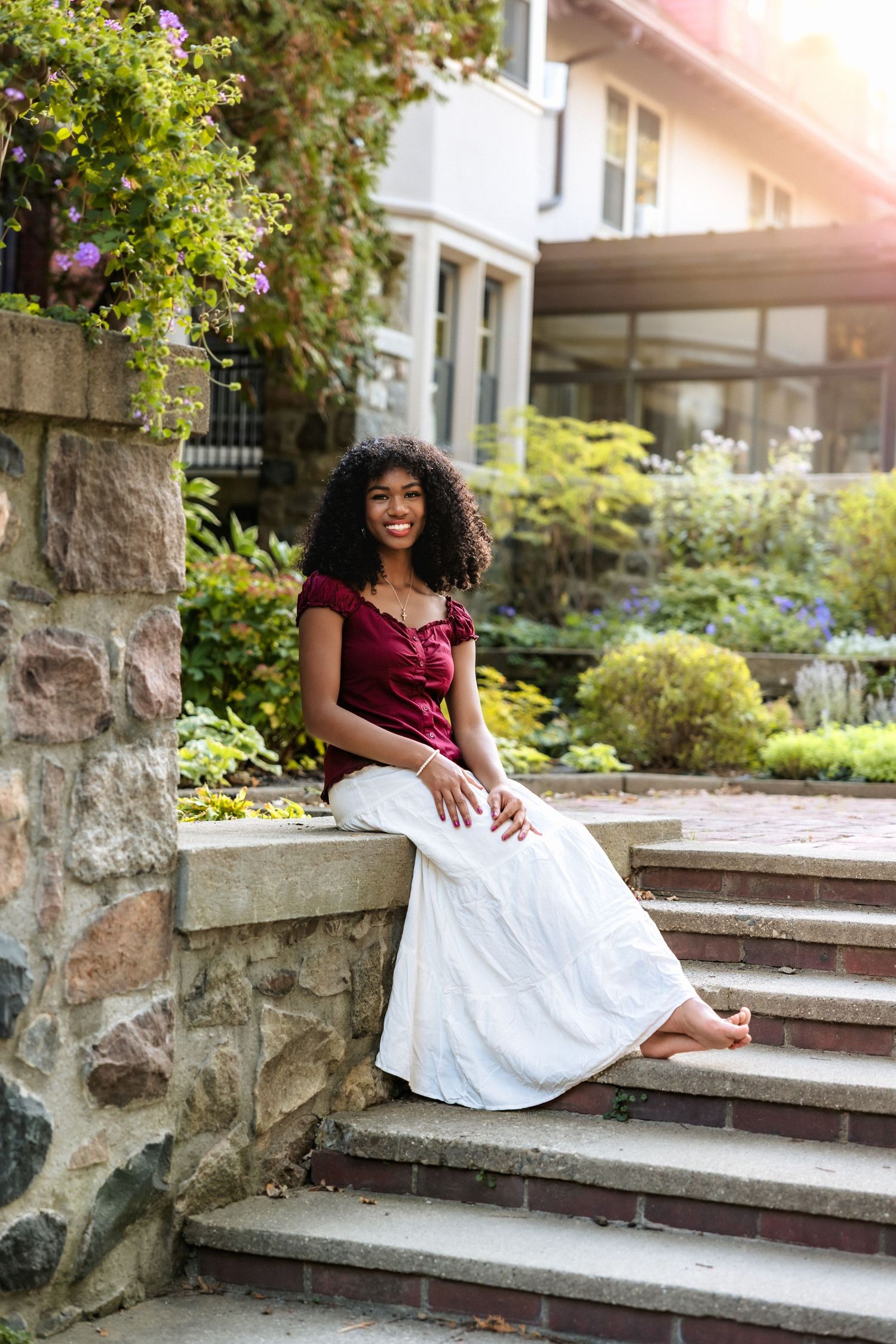 A person sits on stone steps wearing a burgundy top and white skirt, with a house and garden in the background.