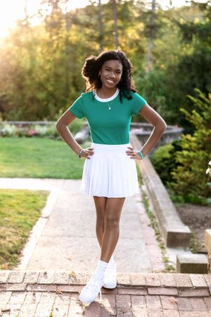 Athletic figure in a green polo shirt and white pleated tennis skirt poses on brick path during golden hour sunset.