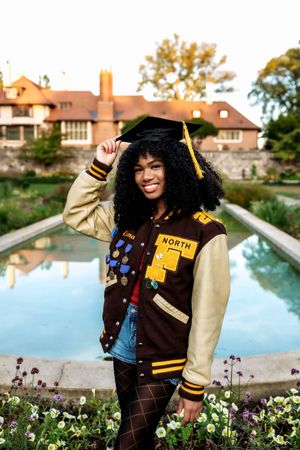 Graduate in varsity letterman jacket and cap poses by reflecting pool with flowers and mansion in background.