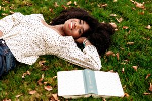 A person in a polka dot dress lies on grass reading a book on a fall day with scattered autumn leaves around them.