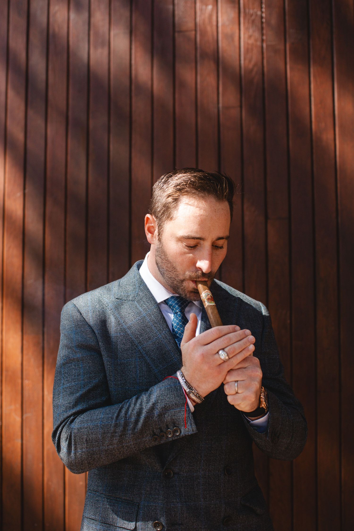 Well-dressed man in gray suit lighting a cigarette against wooden wall background in warm lighting.
