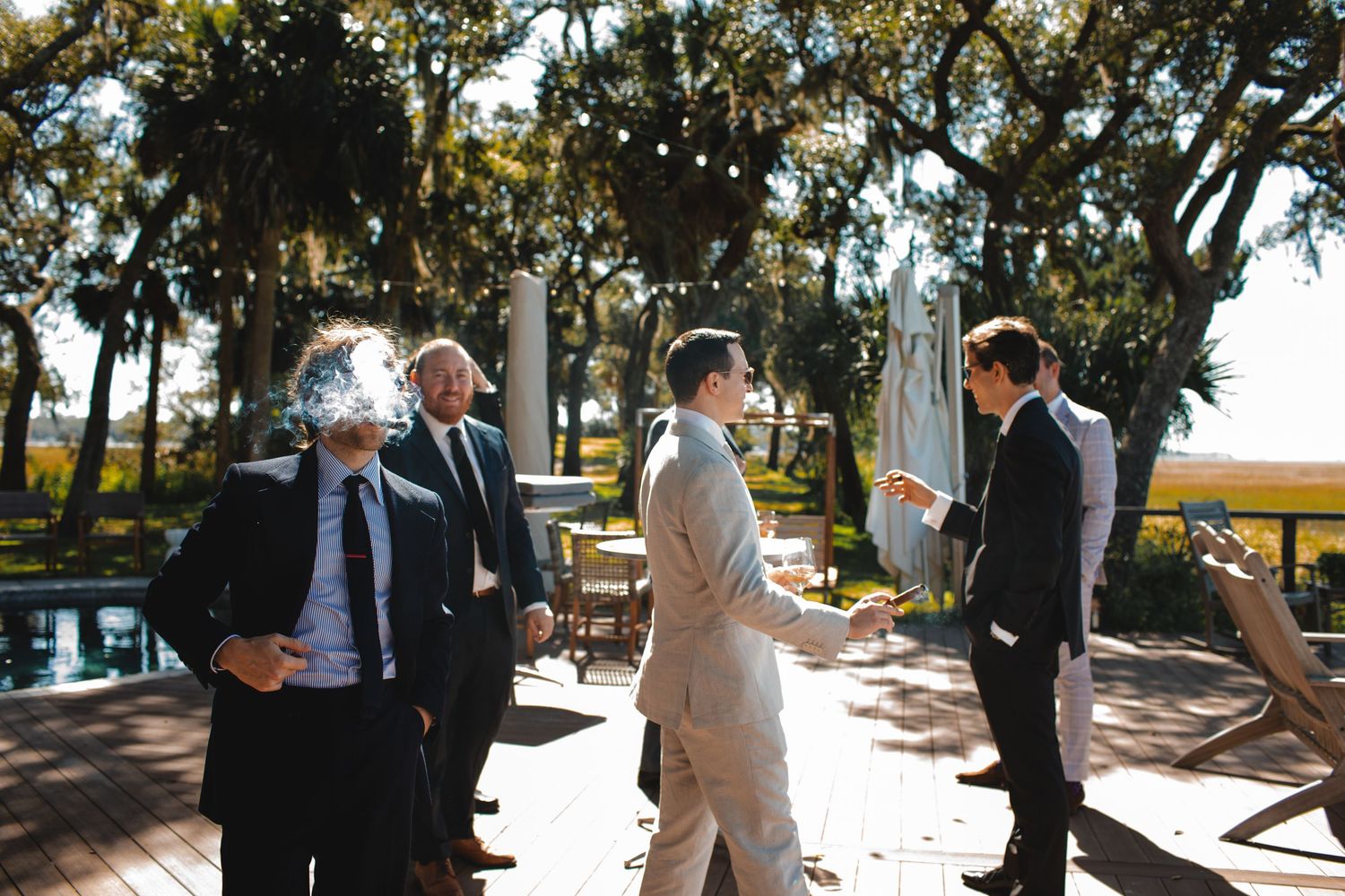 People gathered for an outdoor wedding ceremony on a sunny patio with trees and a waterfront view in the background.