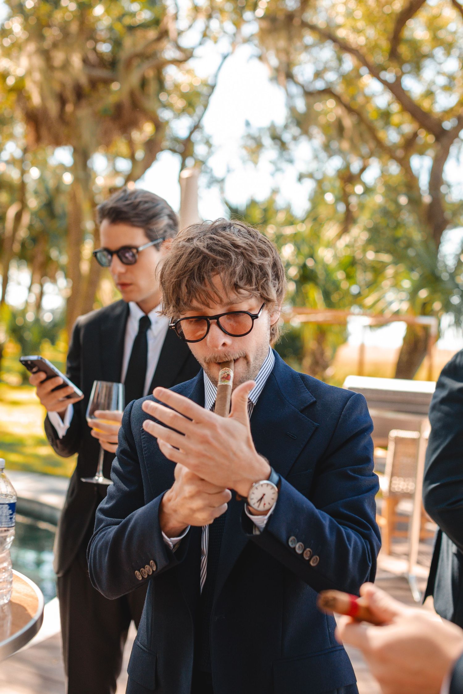 Guests in formal attire lighting cigarettes and checking phones during an outdoor event.