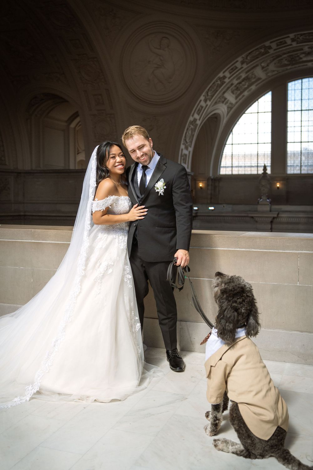 Wedding couple poses with their dog in an elegant marble hallway with ornate architectural details.