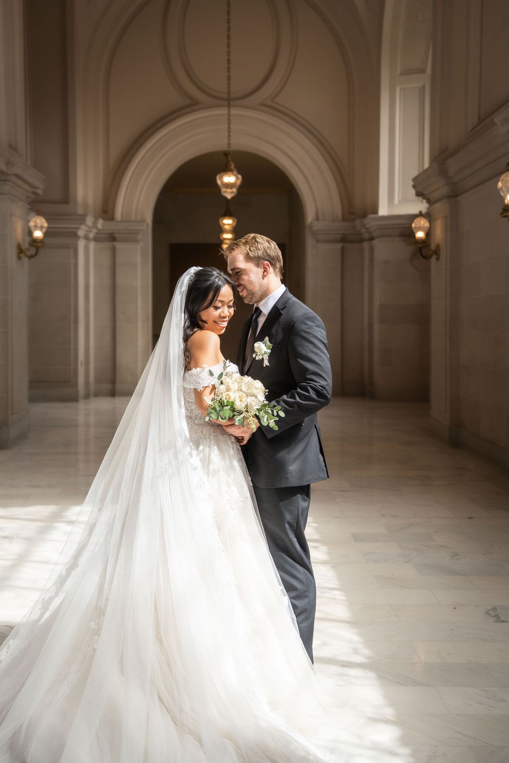 Elegant wedding couple shares intimate moments in a grand marble hallway with natural light streaming through windows.