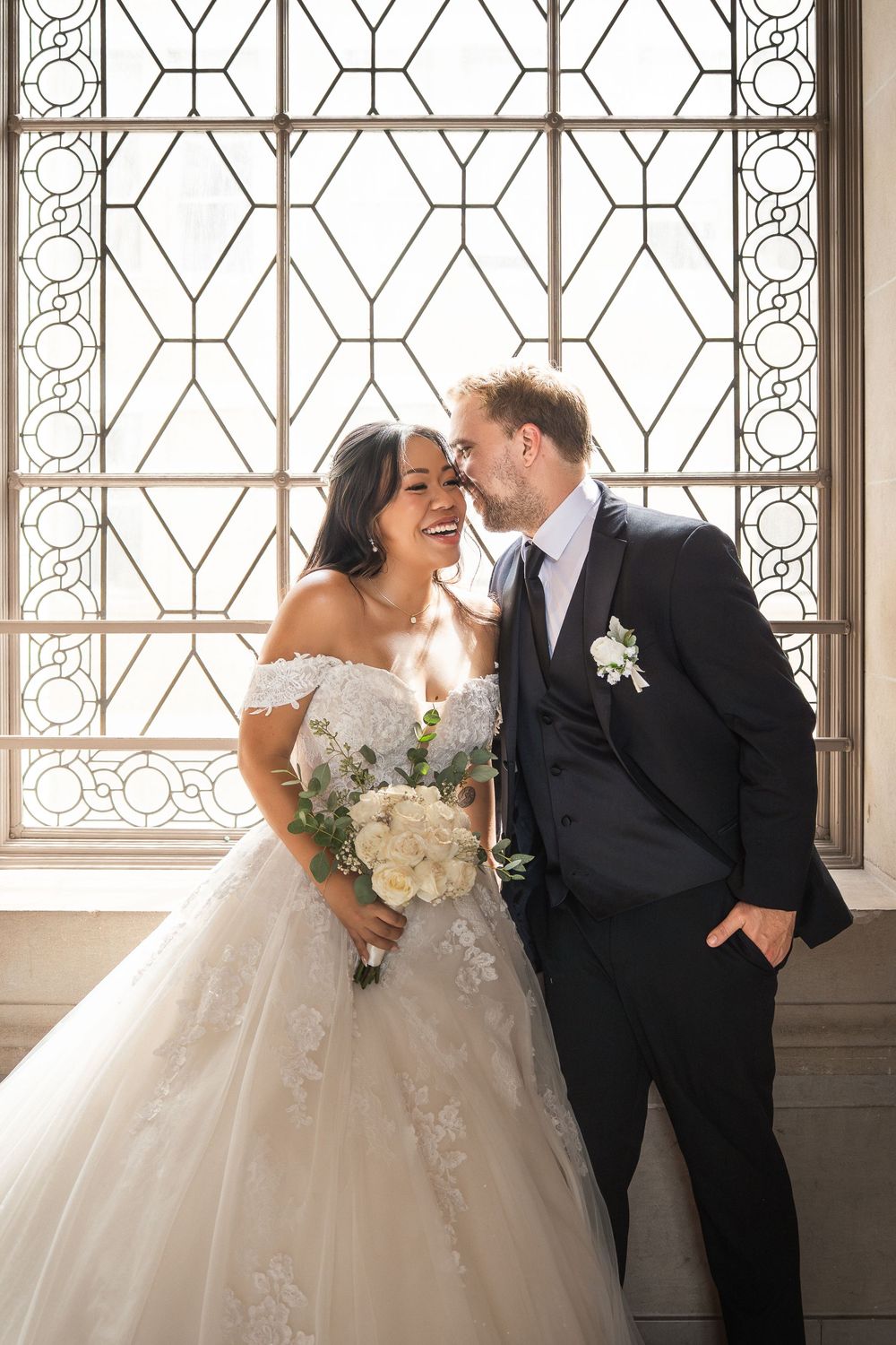 Romantic wedding couple shares intimate moments by ornate window in classic venue with white floral bouquet.