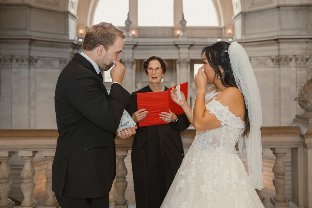 A wedding couple and officiant share a joyful moment during their ceremony in an elegant white interior venue.