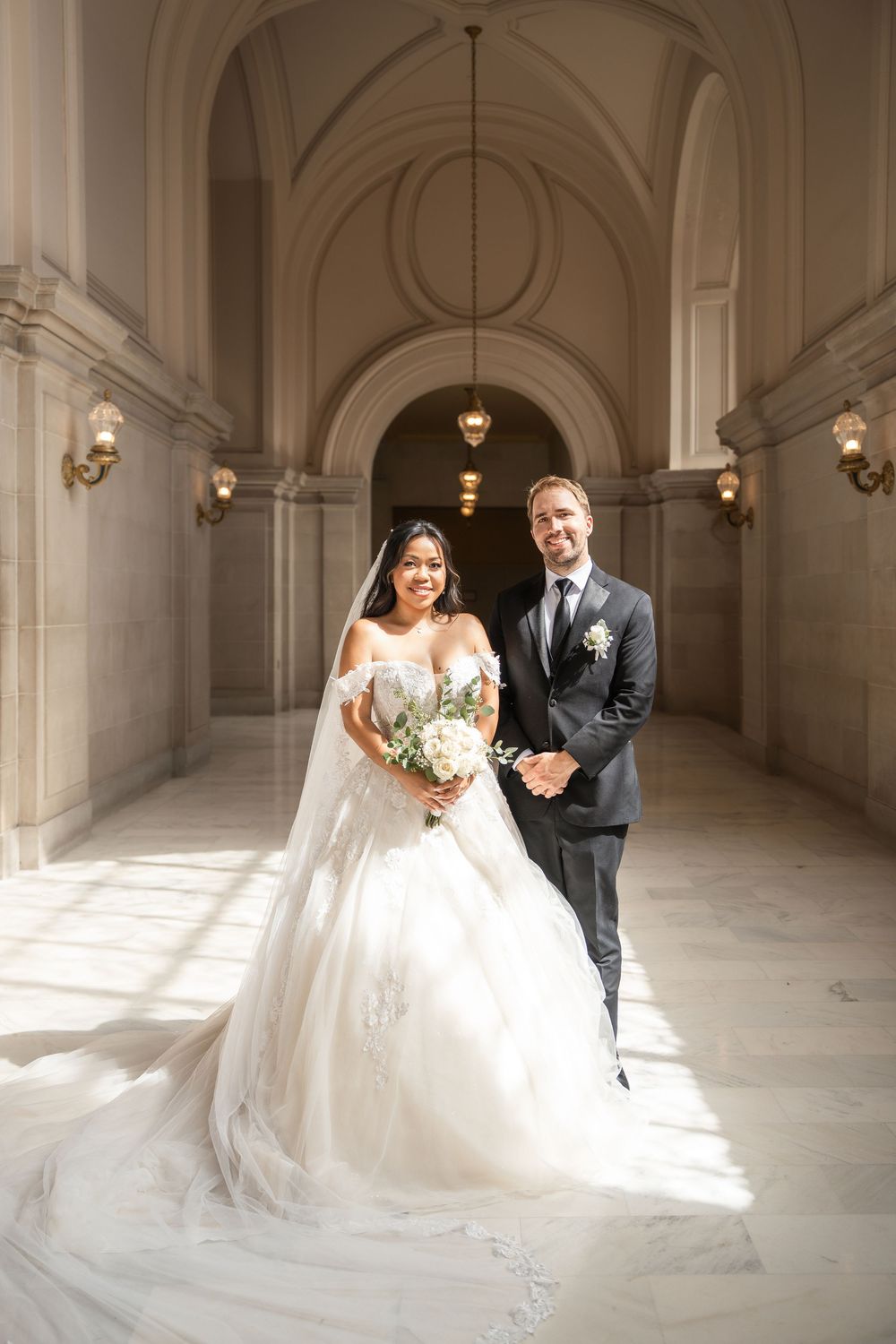 Newlyweds share romantic moment in sunlit marble corridor with grand arched ceilings.
