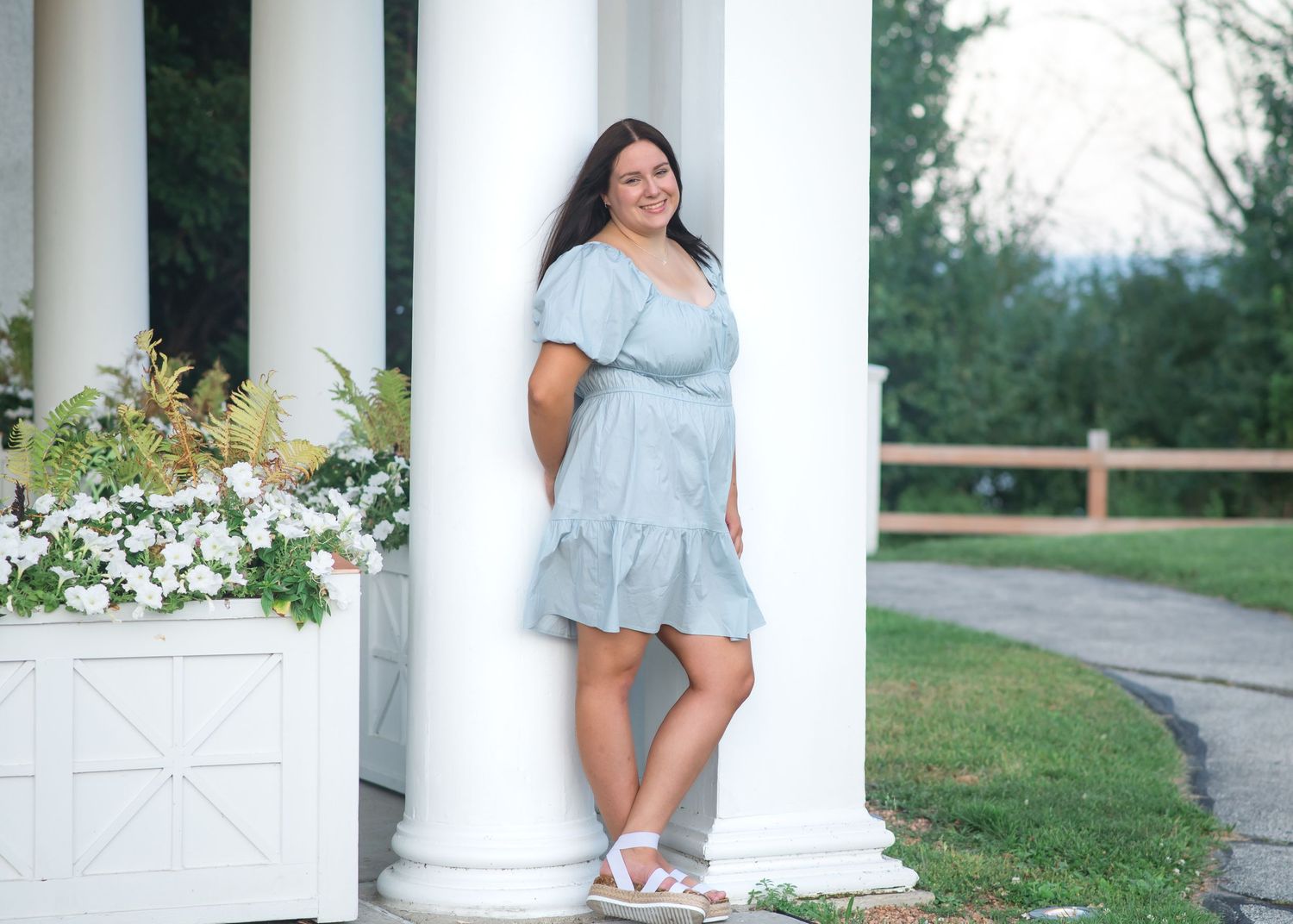 A woman in a light blue off-shoulder dress poses elegantly against white columns in an outdoor garden setting.