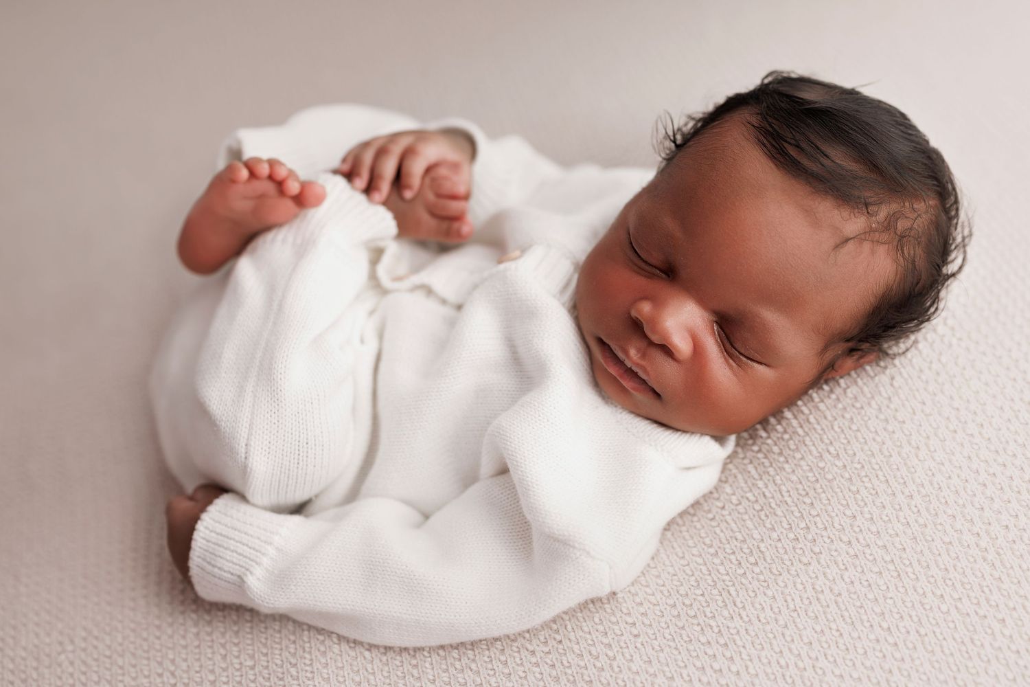 Newborn baby wearing white sleeper outfit lying on textured white blanket in studio setting.