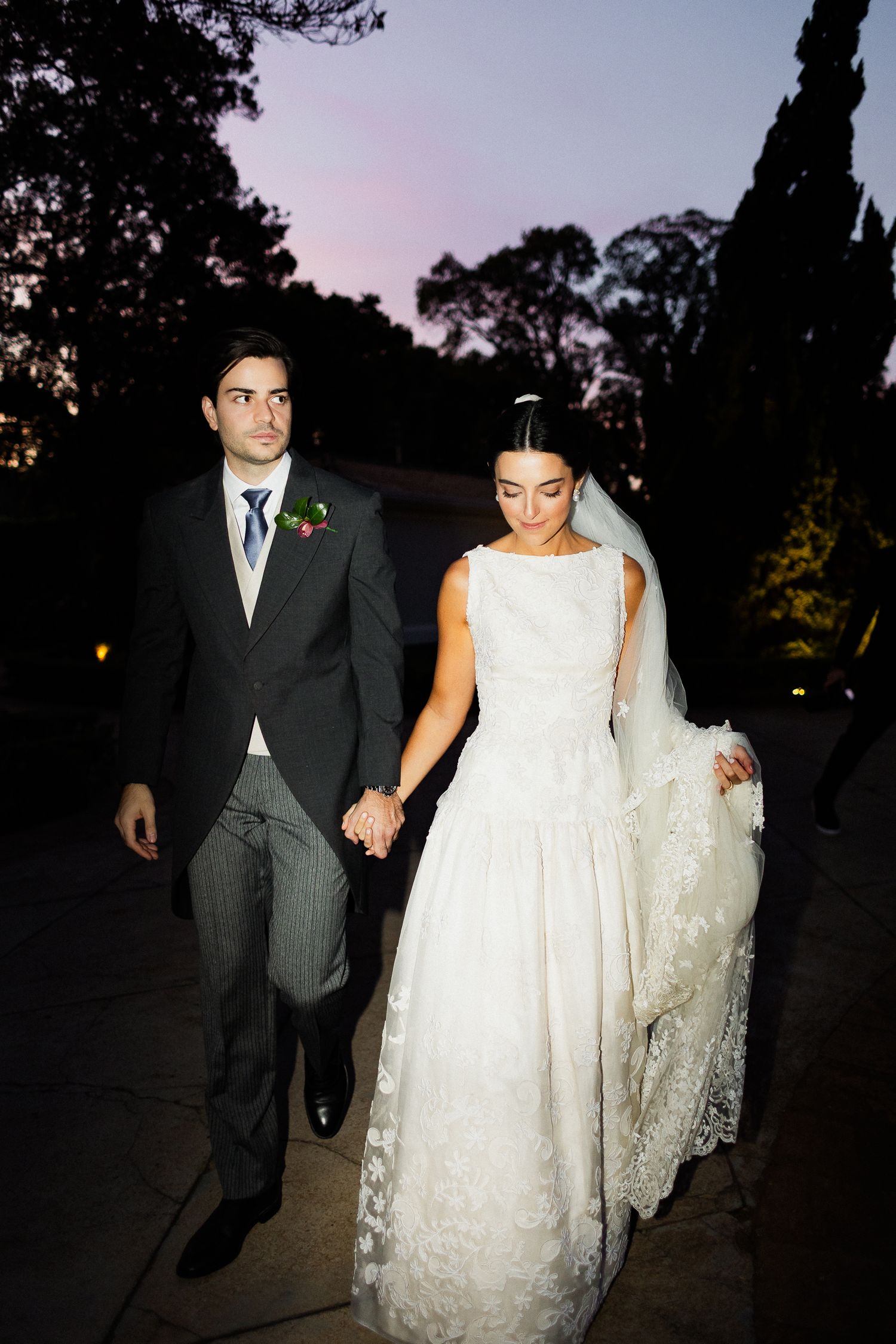 A couple walks together at dusk, with the bride wearing a classic white lace gown and veil while holding her dress.
