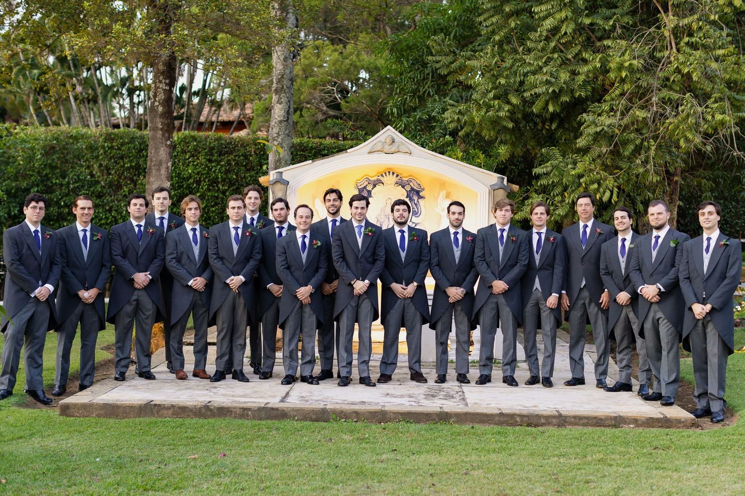 A large group of people in matching navy suits stand in a curved formation on a grassy lawn near trees.