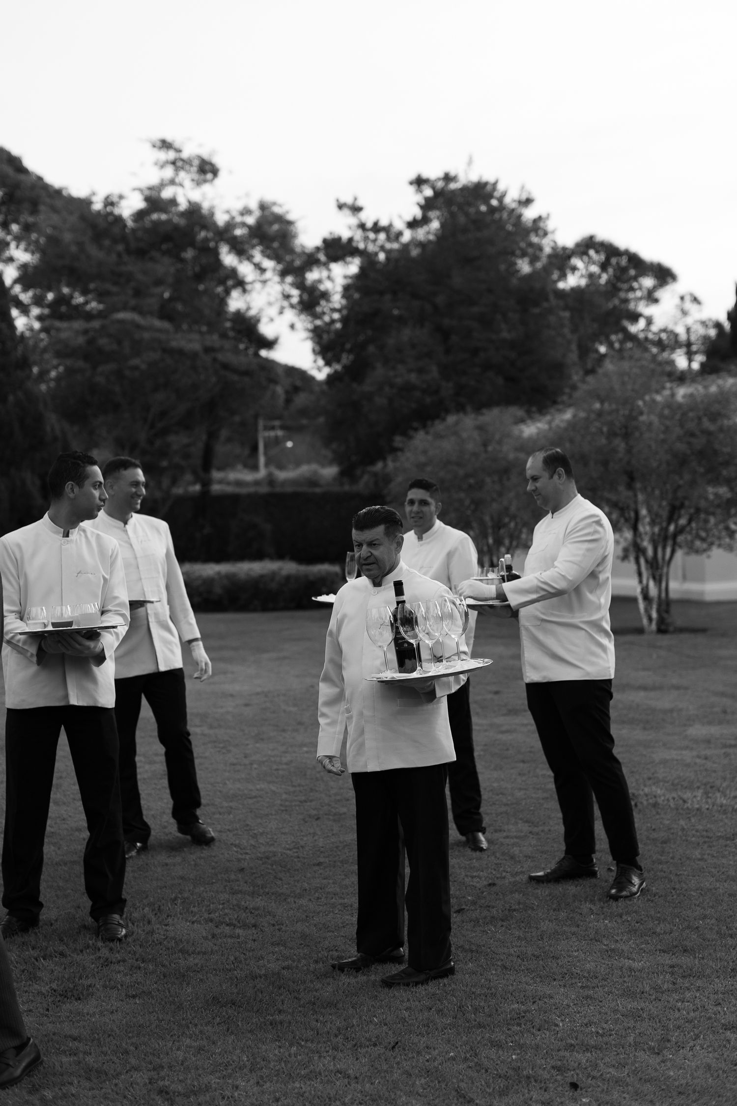 Waiters in white uniforms stand on a lawn carrying serving trays in this vintage black and white photograph.