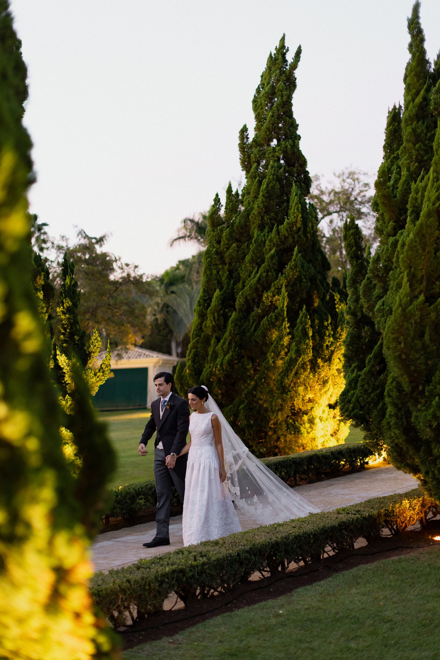 A couple walks along a garden path lined with tall cypress trees at sunset with warm lighting.