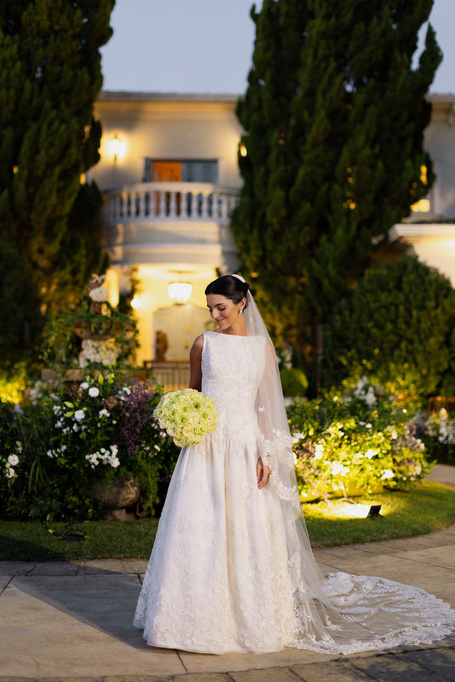 A bride in an elegant white lace wedding gown poses in a garden setting with illuminated cypress trees at dusk.